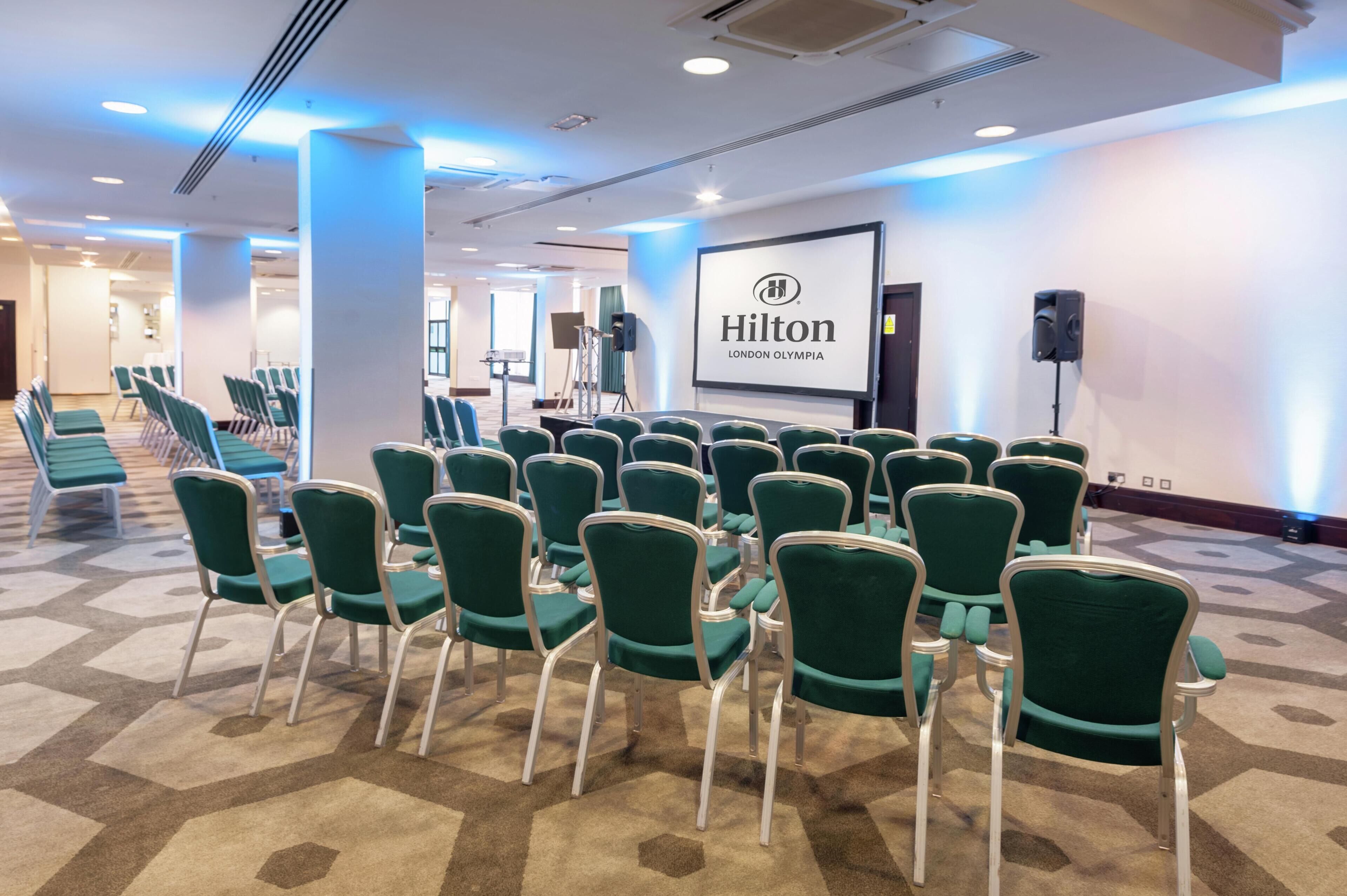 A conference room with green chairs arranged in rows, a projector screen with the Hilton logo, and a speaker on a stand.