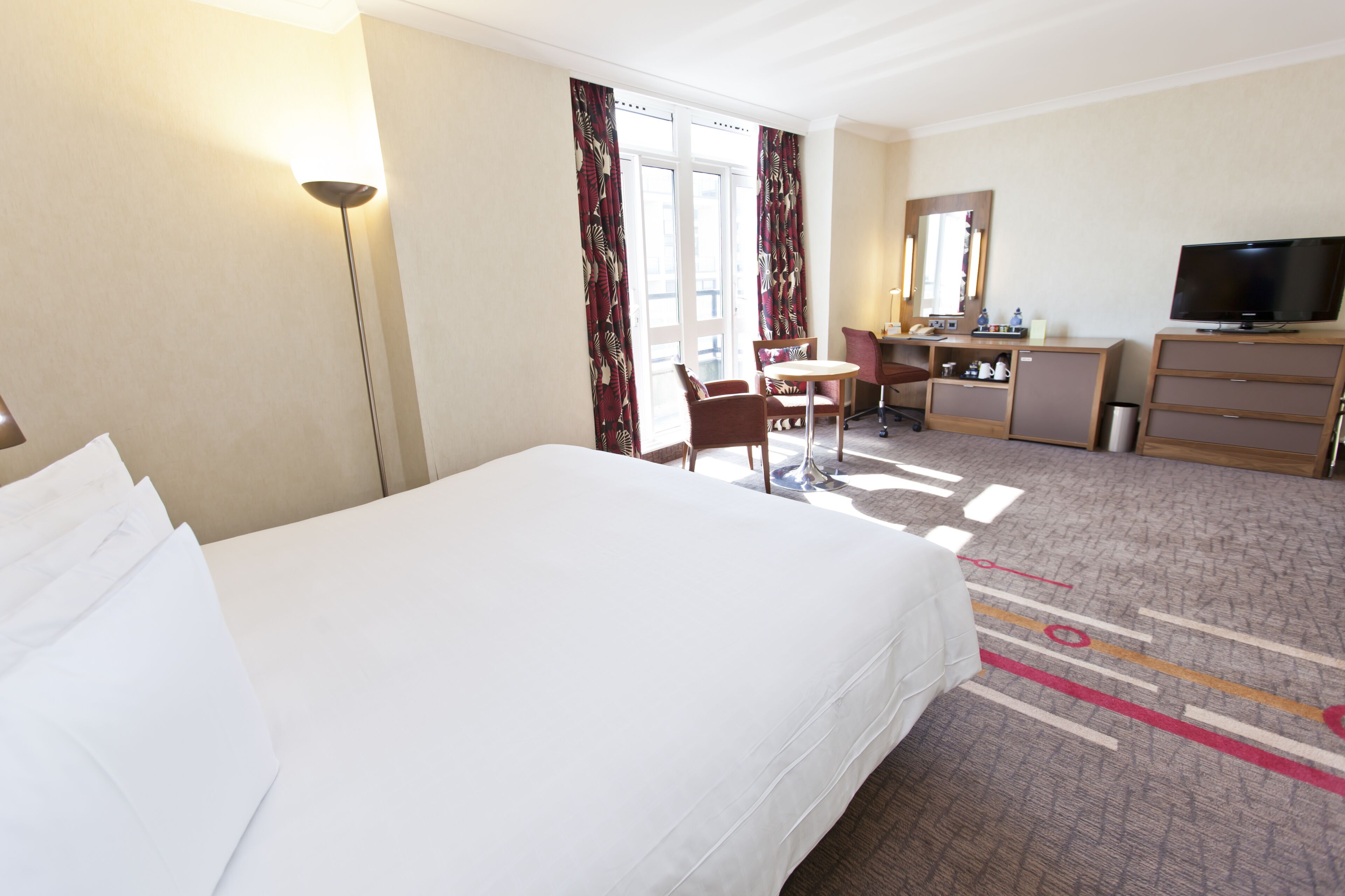 A hotel room with a large bed, a desk with a chair, a television, and a small cabinet.