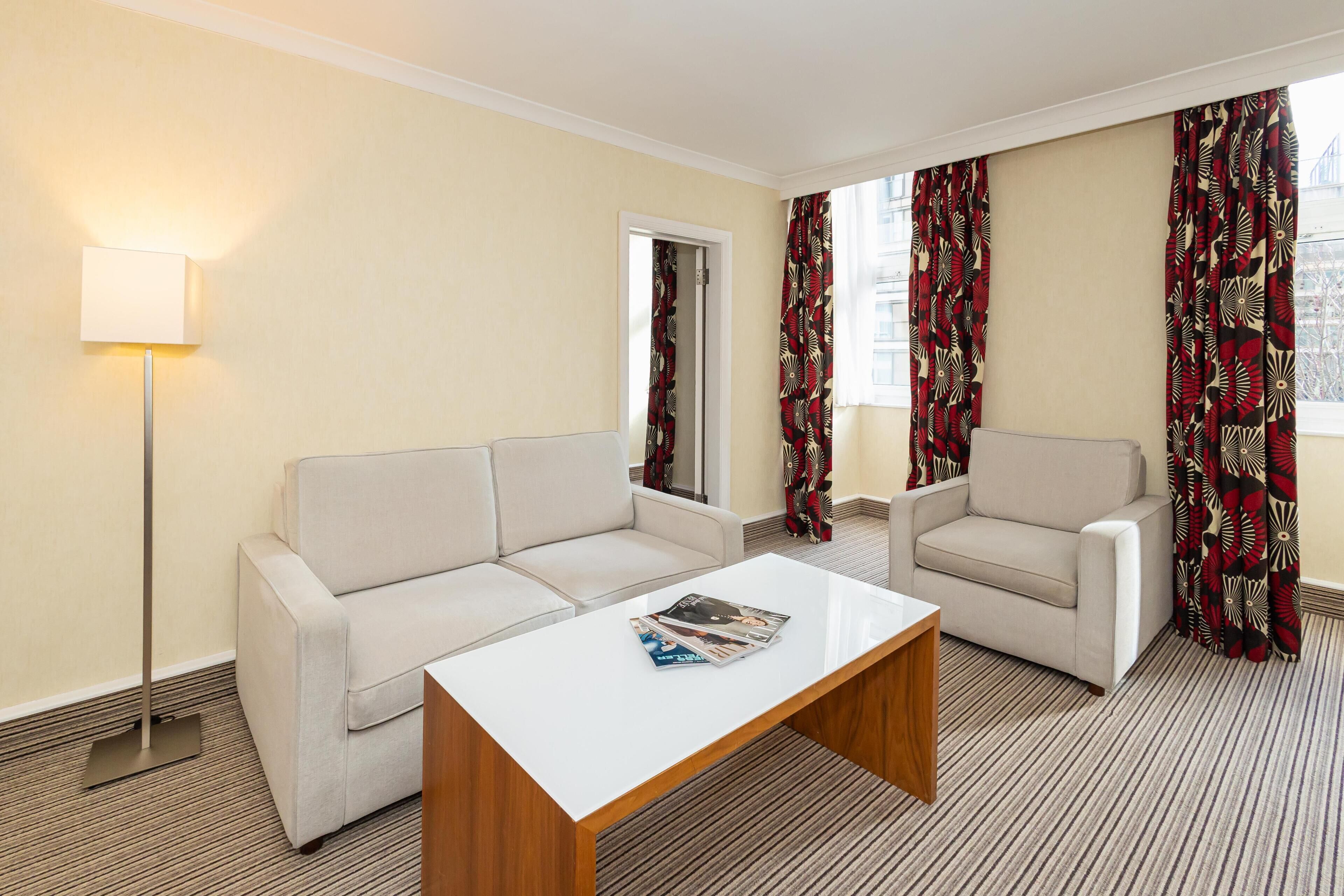 A hotel room with a sofa, a coffee table, a lamp, and patterned curtains.
