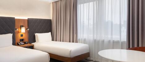 A hotel room with two beds, a bedside table with a lamp, a telephone, and a view of the city through sheer curtains.