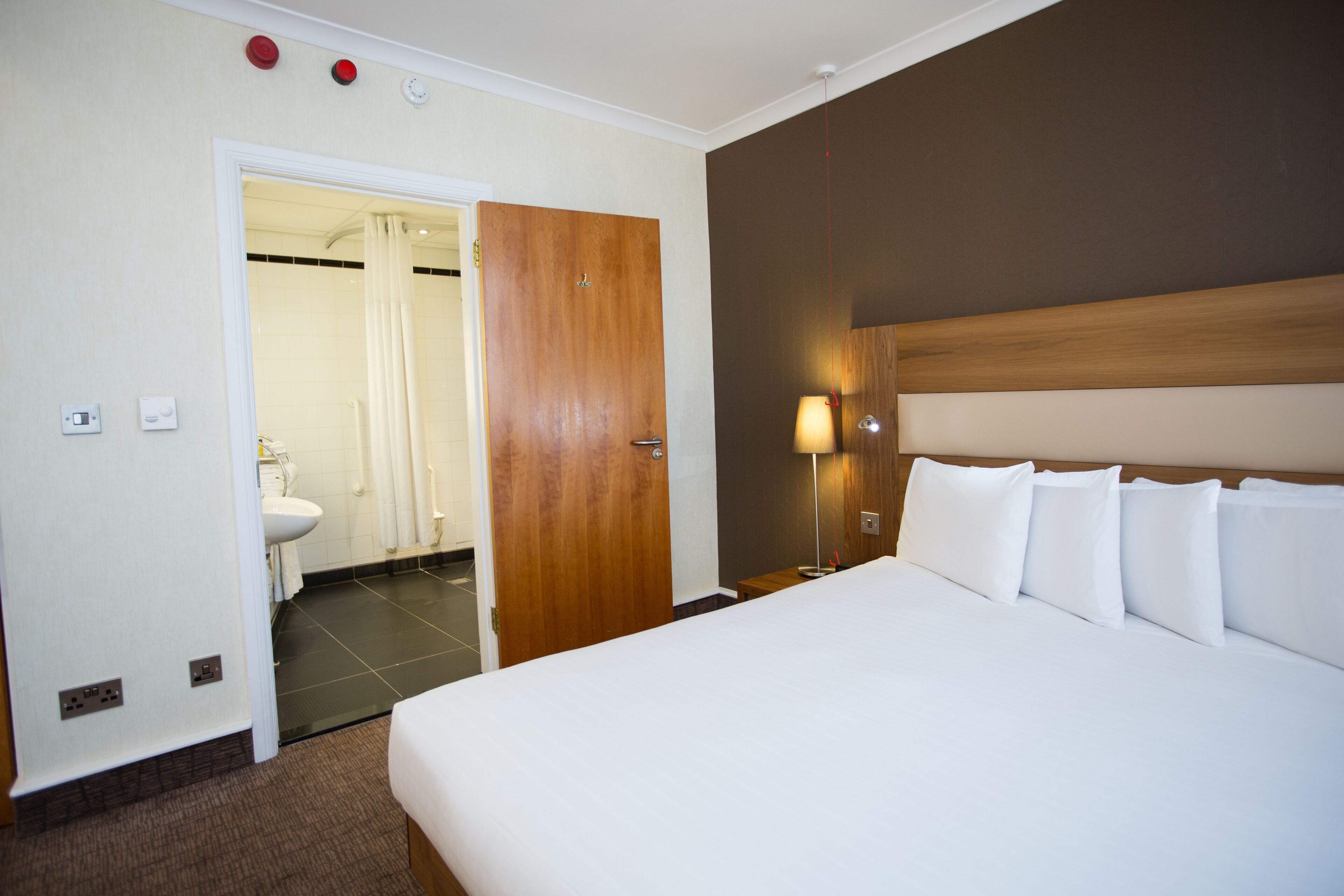 A hotel room with a large bed, a wooden headboard, a bedside table, a lamp, and a bathroom visible through an open door.