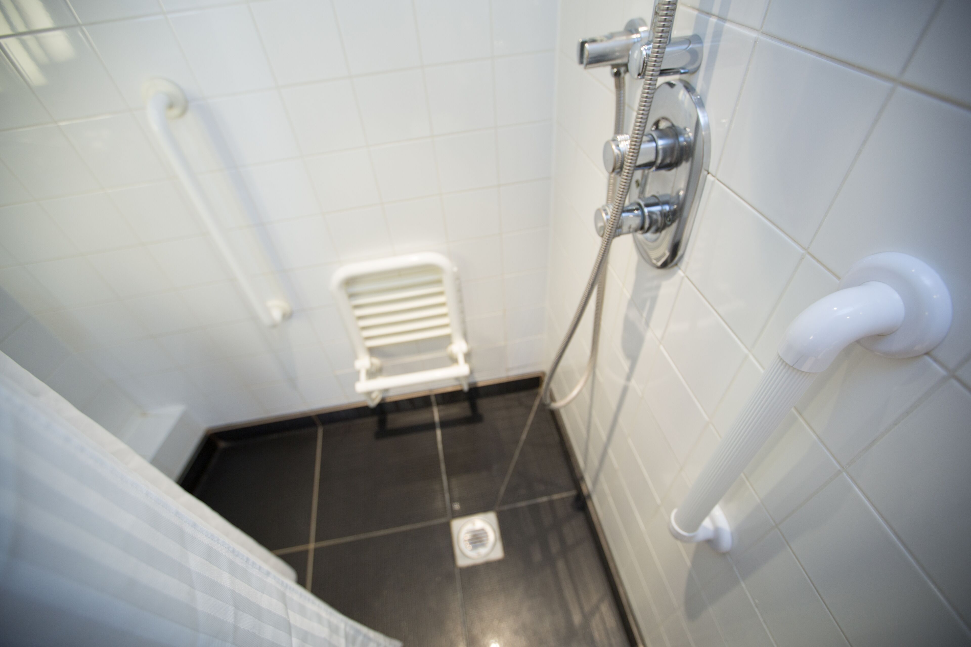 Une salle de bain avec des murs recouverts de carreaux blancs, un sol noir, des toilettes blanches et une douche avec un flexible.
