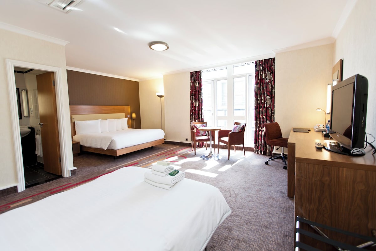 A hotel room with a large bed, a desk with a computer, a chair, and a small table with a lamp.
