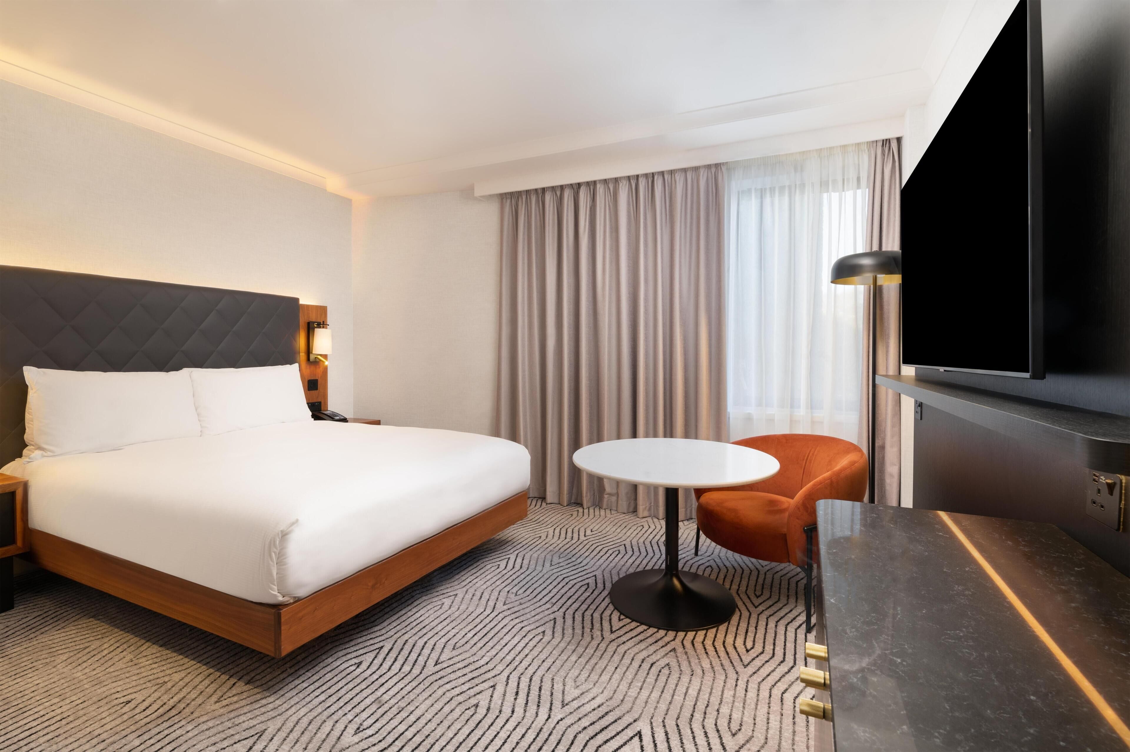 Habitación de hotel con cama, una mesa redonda, una silla naranja, televisor y ventana con cortinas.