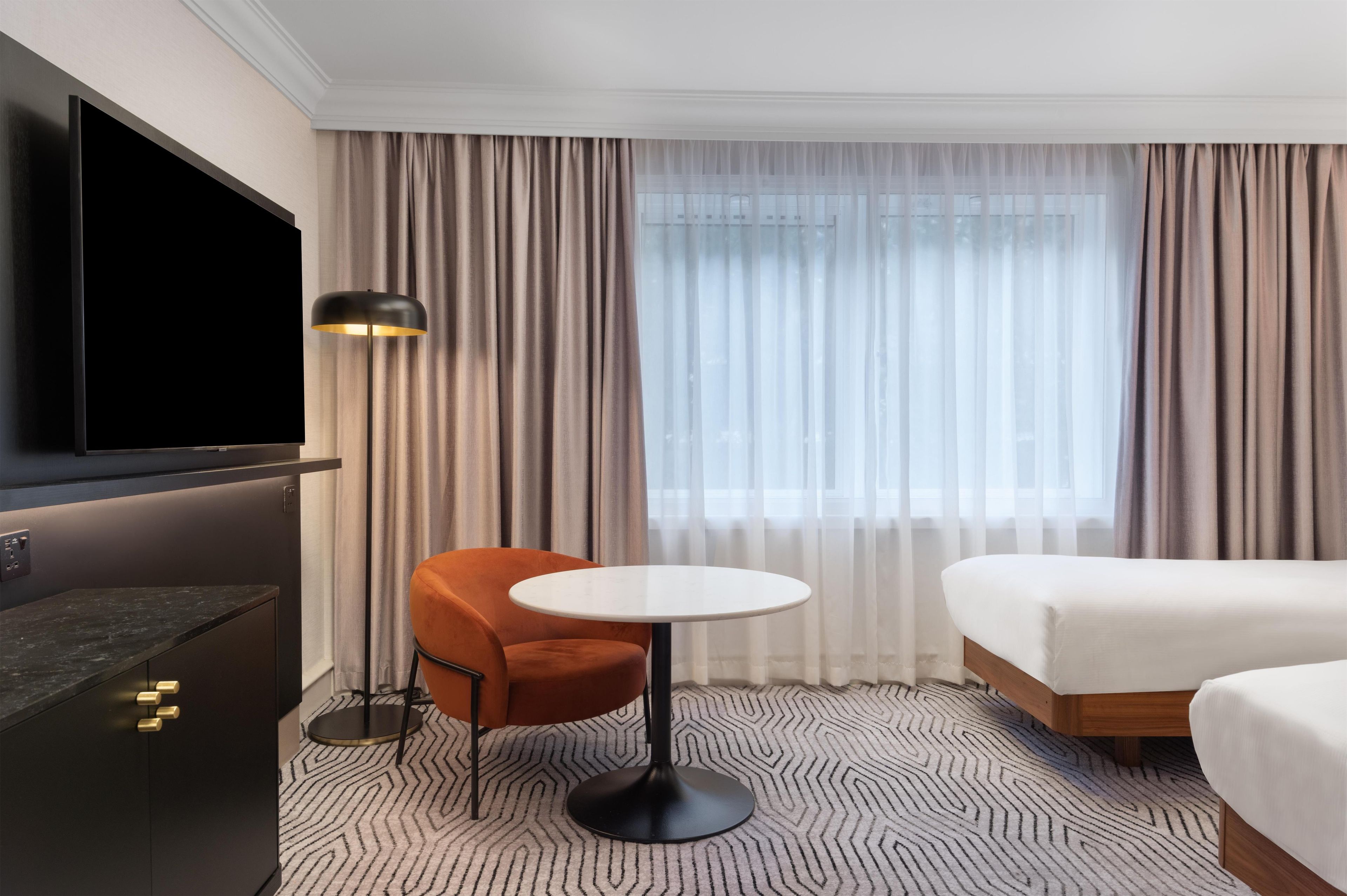 Une chambre d’hôtel comprenant un lit, un bureau, une chaise, une table ronde, un téléviseur et une fenêtre avec des rideaux.