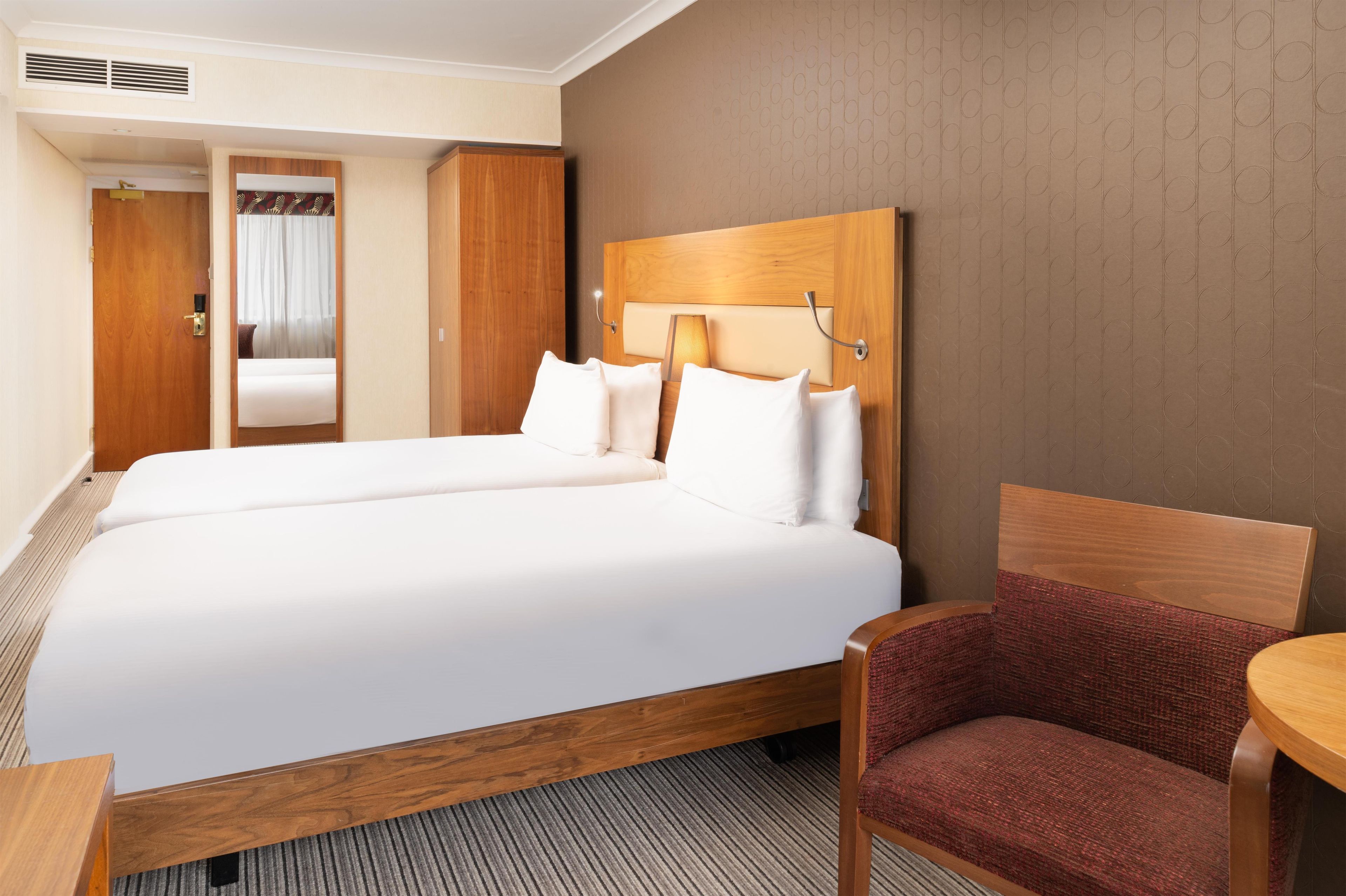 A hotel room with a double bed, a wooden headboard, a small table, a chair, and a door leading to a bathroom.