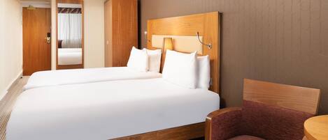 A hotel room with a double bed, a wooden headboard, a small table, a chair, and a door leading to a bathroom.