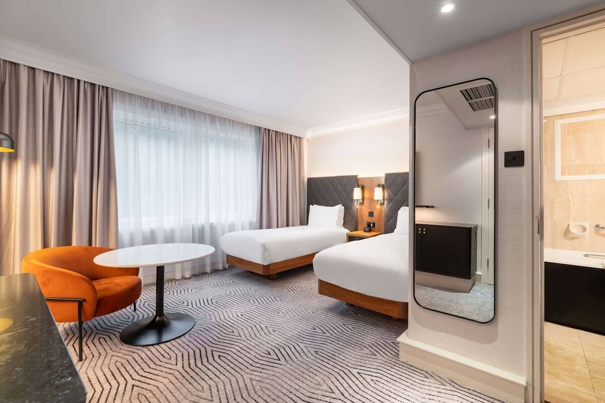 A hotel room with two beds, a small table, an orange chair, a large mirror, and a television.