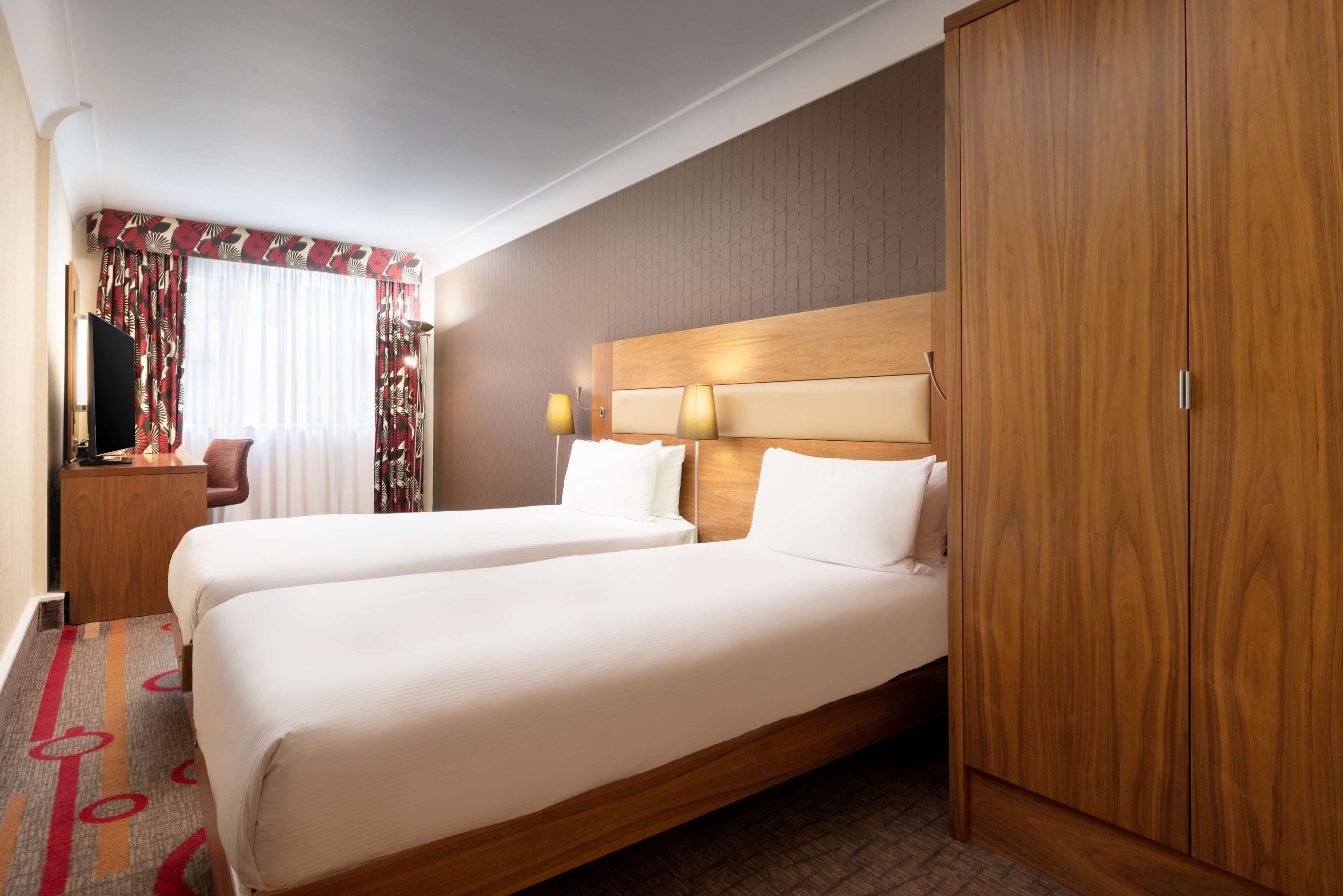 A hotel room with two beds, a wooden wardrobe, a desk with a lamp, and a window with curtains.