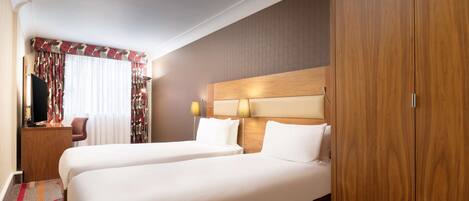 A hotel room with two beds, a wooden wardrobe, a desk with a lamp, and a window with curtains.