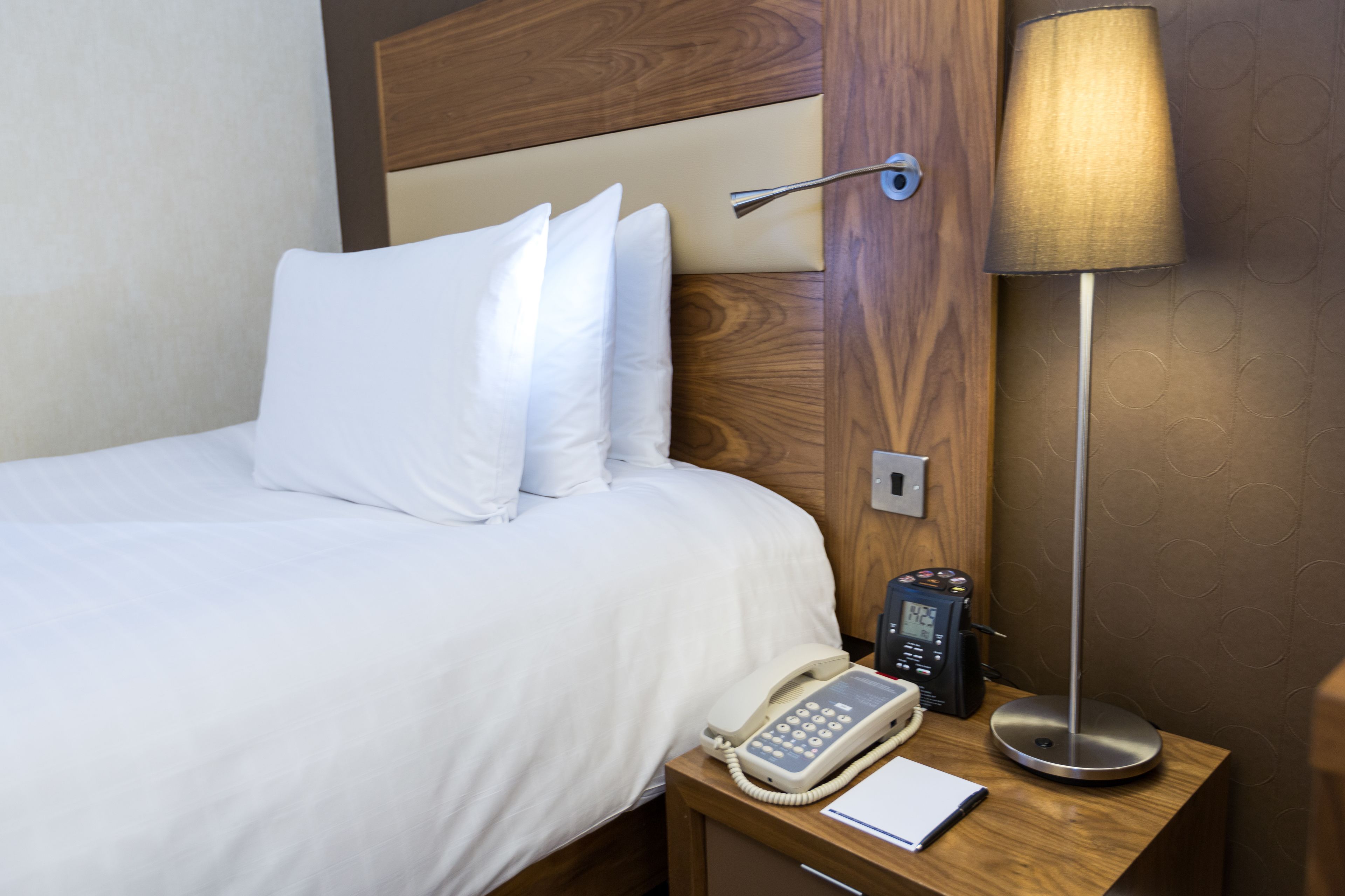 A hotel room with a bed, a bedside table with a lamp, a phone, and a notepad.