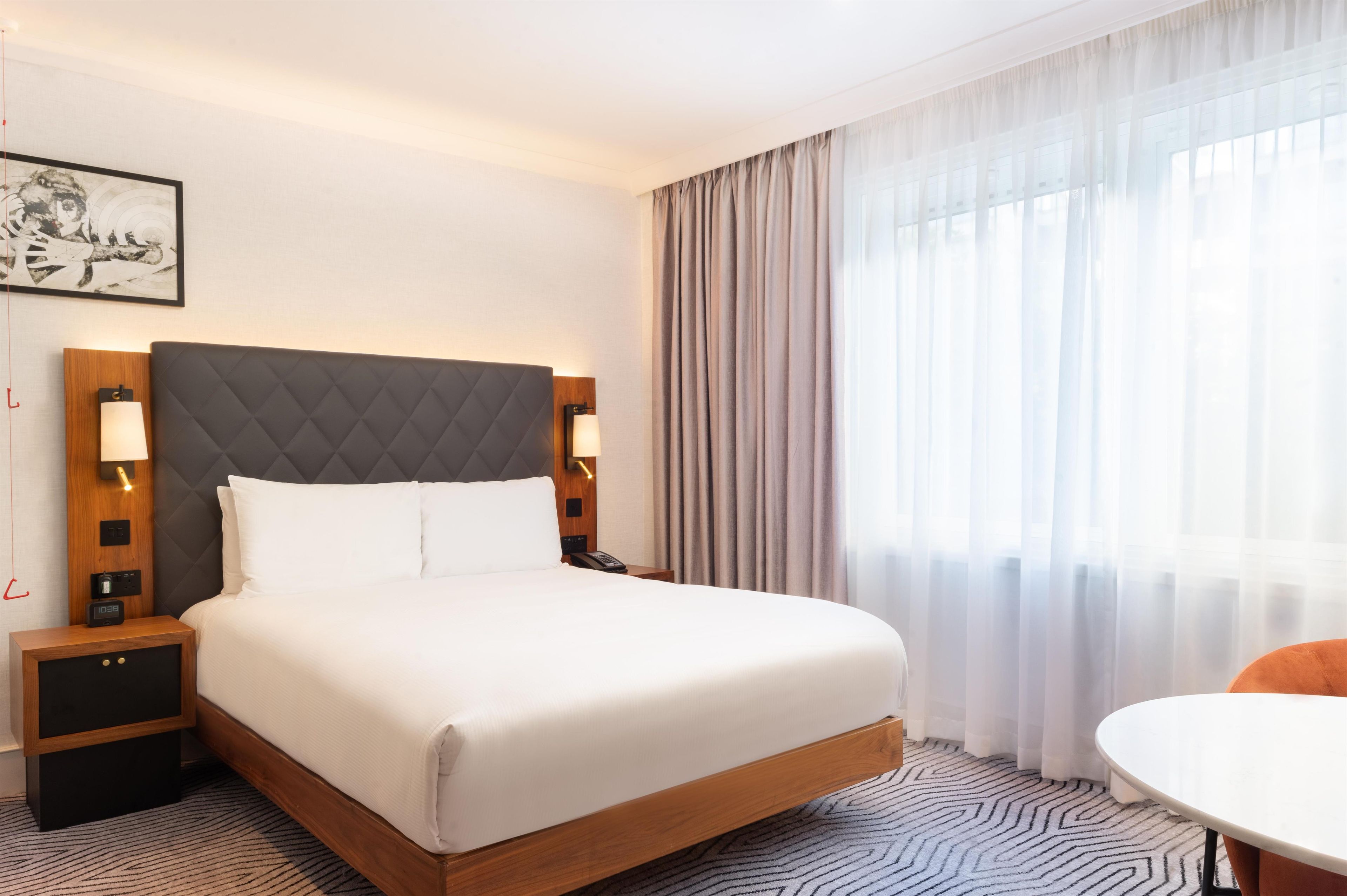 Une chambre d’hôtel avec un lit, des lampes de chevet, une horloge, un téléphone, un tableau au mur et une fenêtre avec des rideaux.