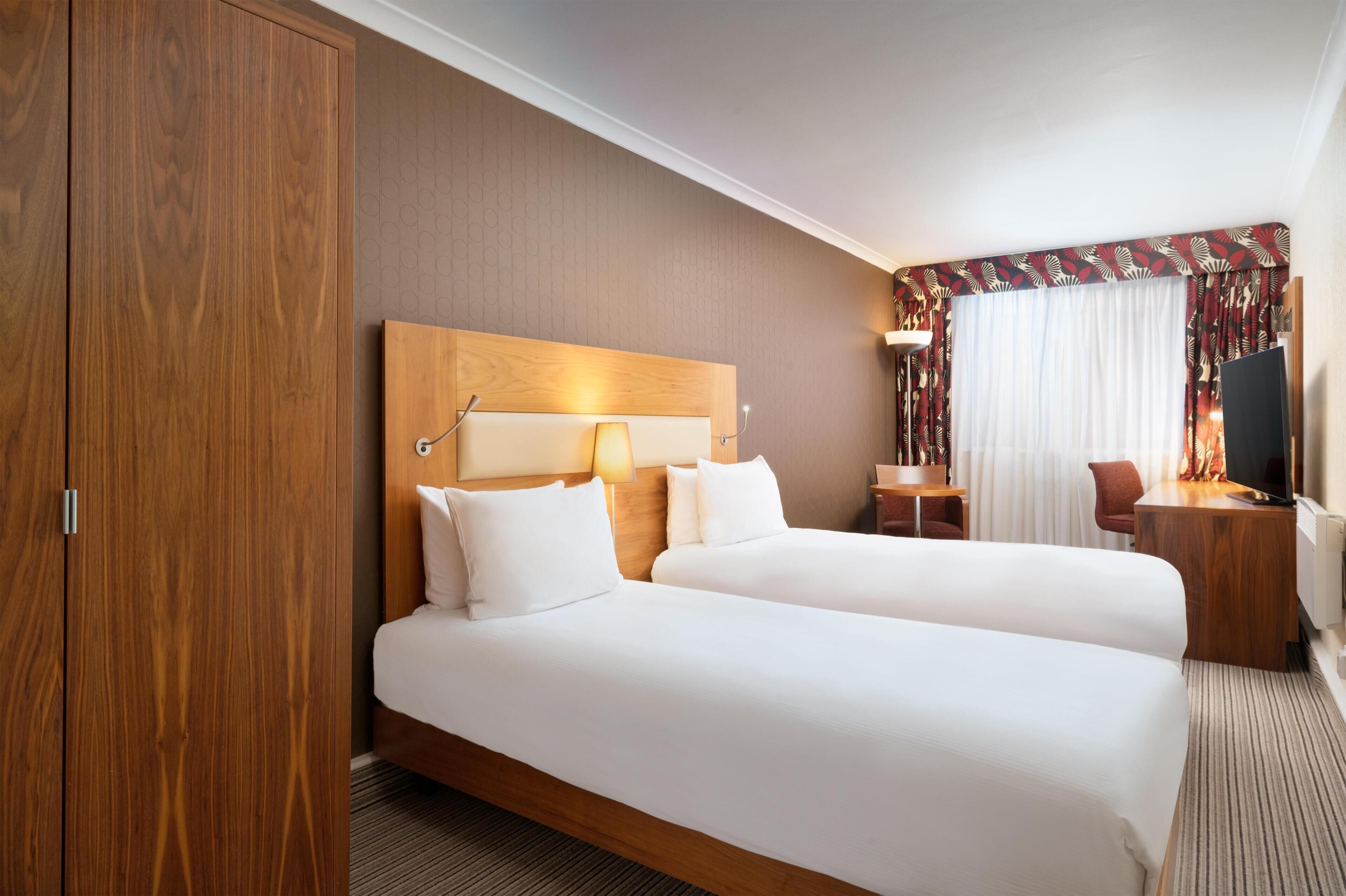 A hotel room with two beds, a wooden wardrobe, a desk with a chair, a TV, and a window with curtains.