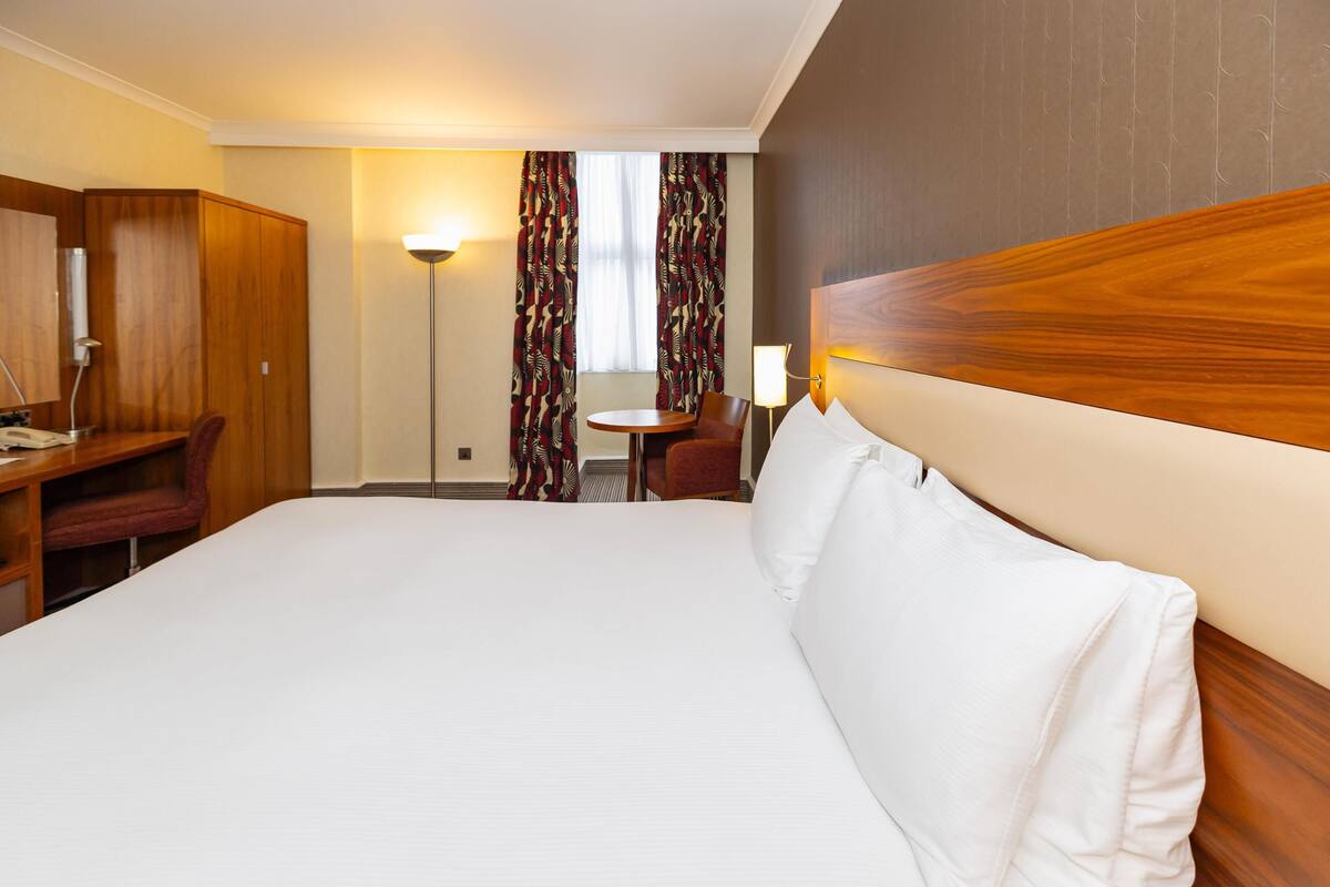 A hotel room with a large bed, a desk with a chair, a small table, and a lamp.