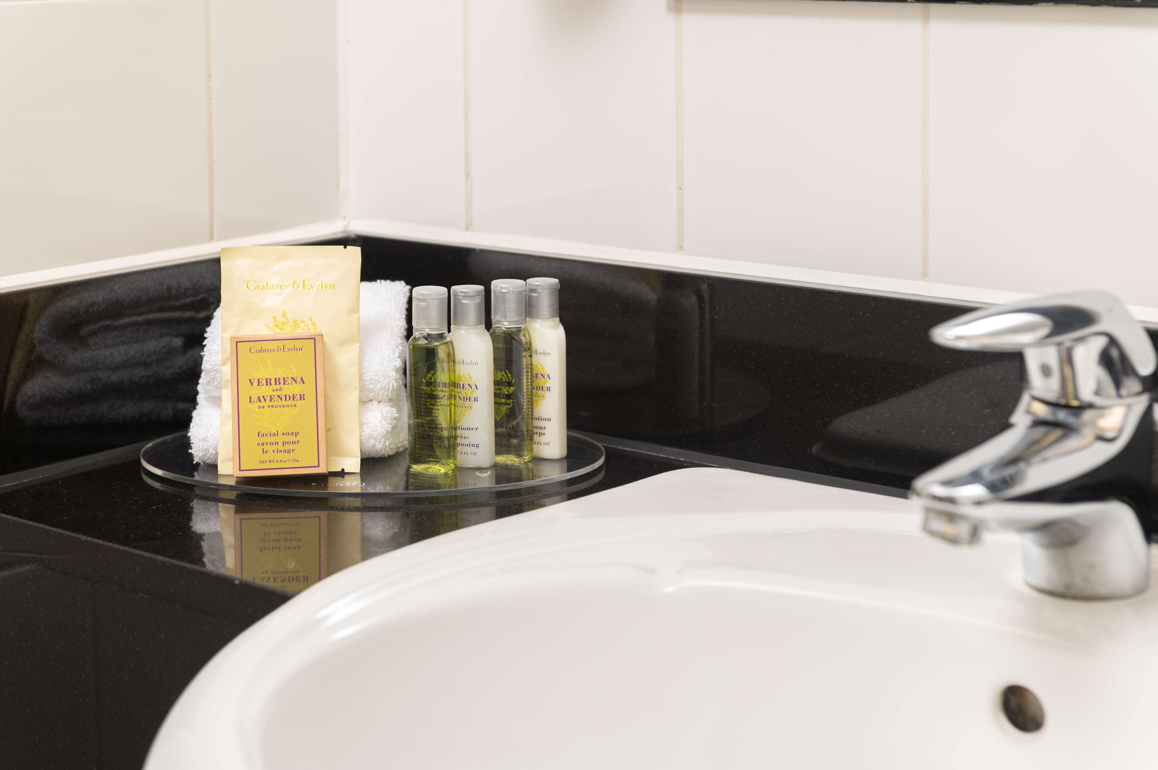 A bathroom sink with a white basin and a chrome faucet. On the counter, there is a package of Verbenal Lavender facial soap and four bottles of lavender lotion.