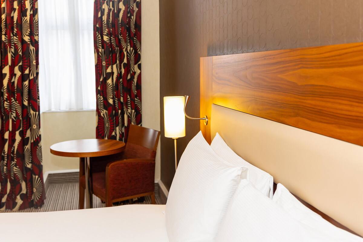 A hotel room with a bed, a round table, a chair, a lamp, and curtains.
