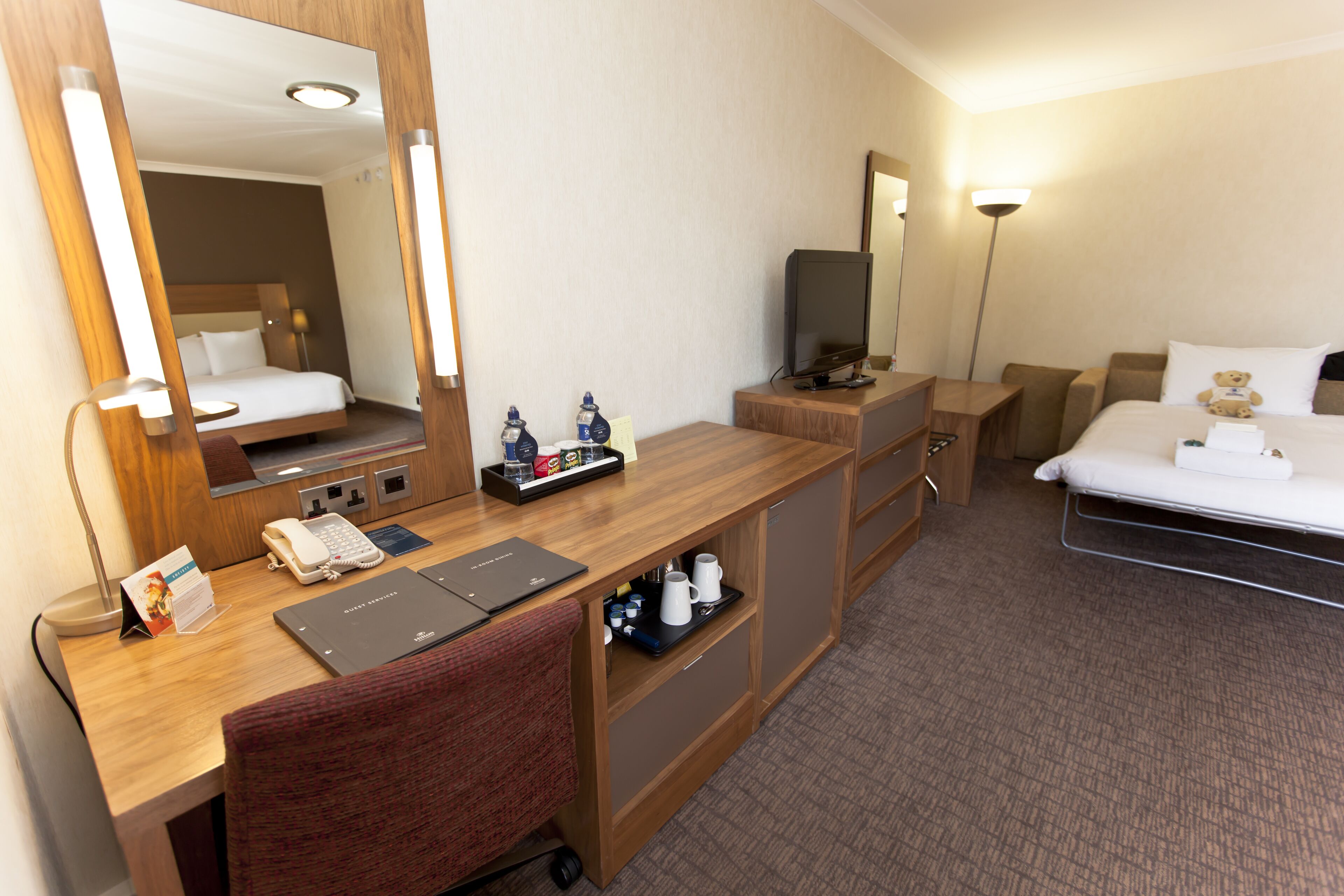 A hotel room with a wooden desk, a bed, a television, and a mirror.