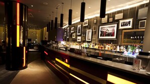A bar area with a long counter, hanging lights, and framed pictures on the wall. - Hilton London Olympia (London)