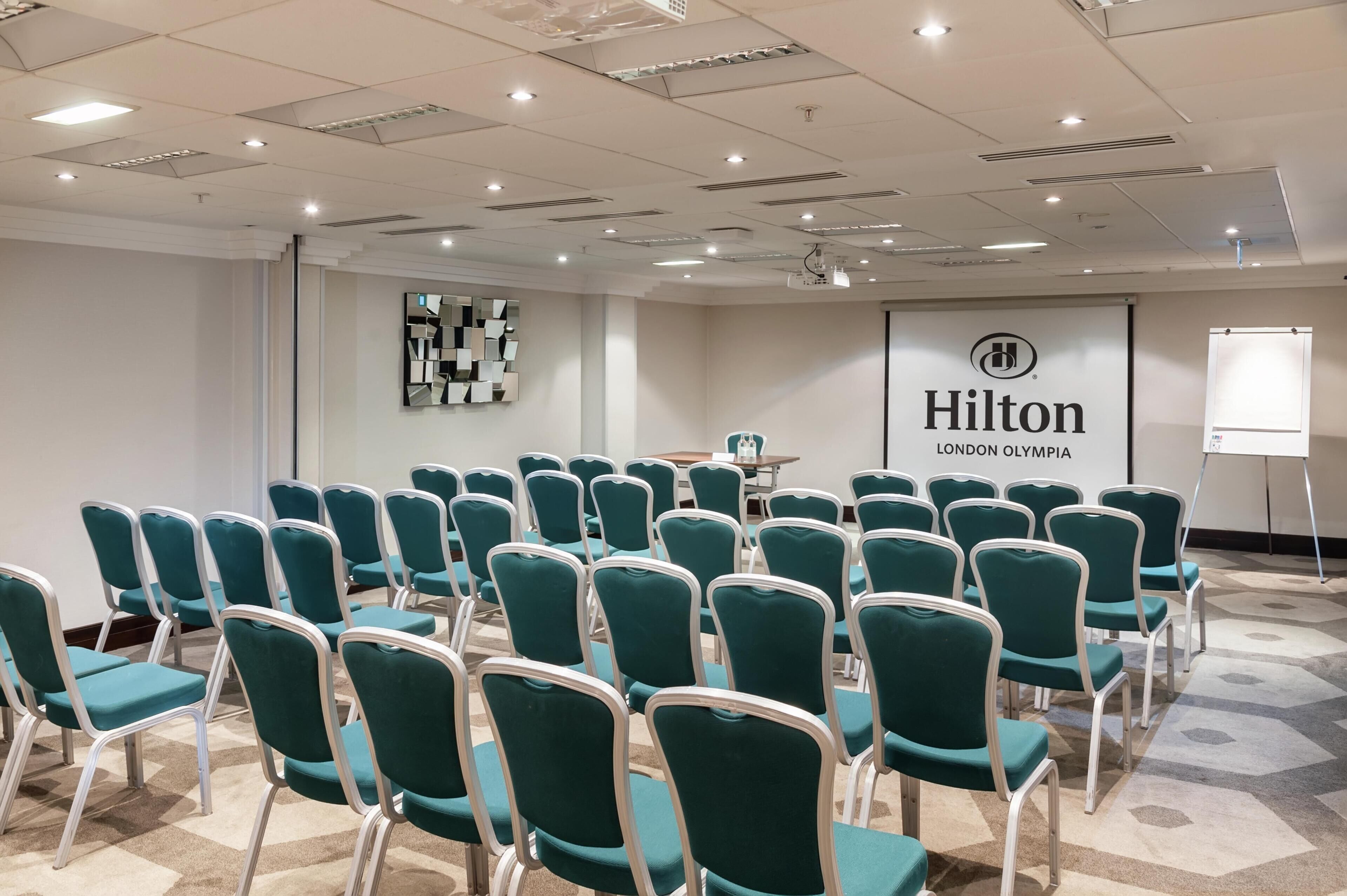 A conference room with rows of teal chairs, a projector screen, and a 'Hilton London Olympia' sign.