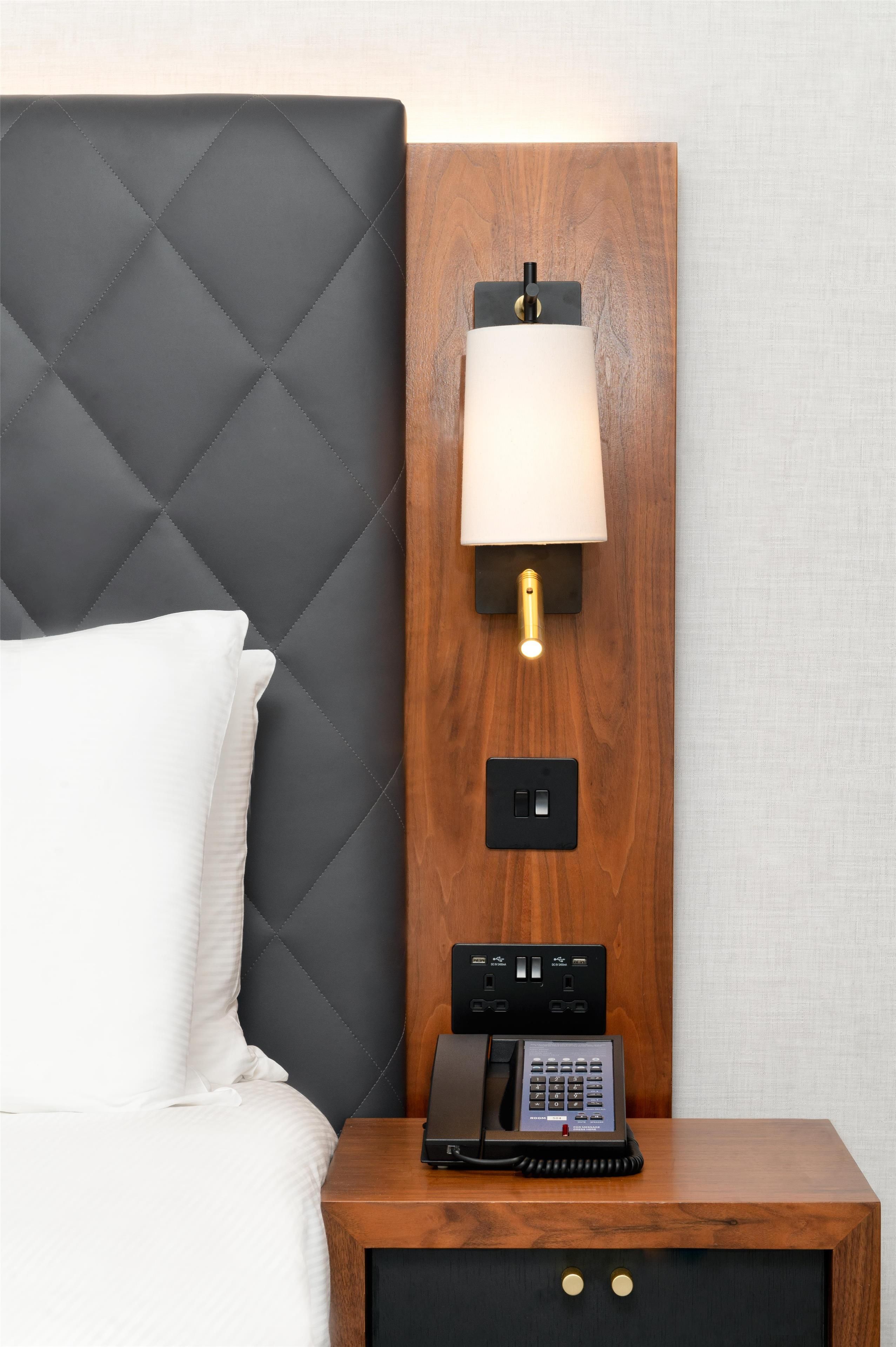 Una cama con cabecera negra, una mesita de noche con una lámpara, un teléfono e un enchufe.
