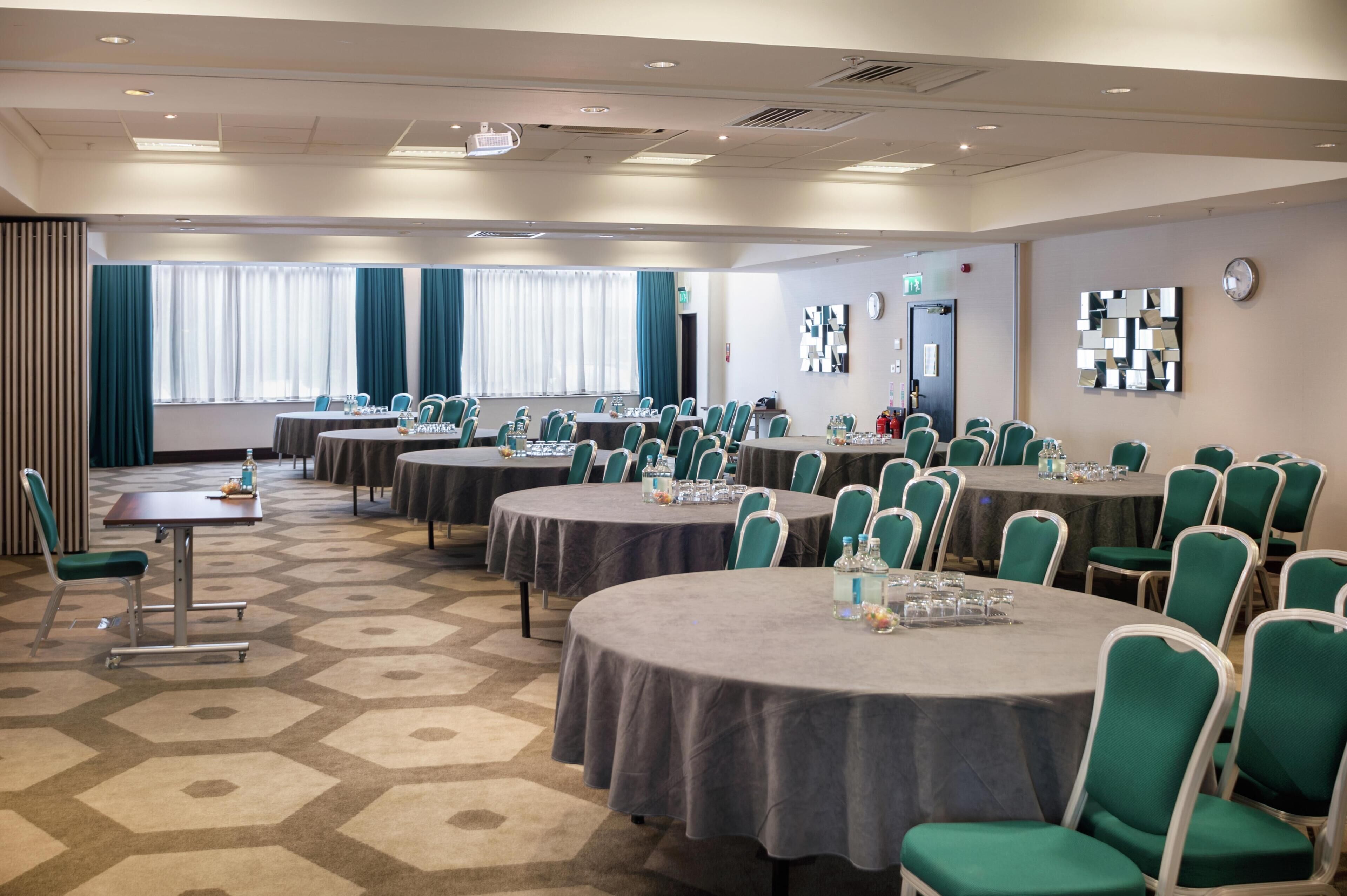 A conference room with multiple tables and chairs, arranged in a U-shape, with a carpeted floor and large windows with curtains.