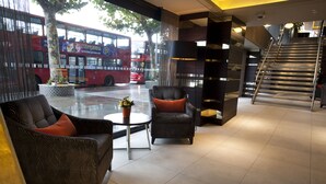 A modern lobby with a sofa, armchair, and small table, a staircase, and a red double-decker bus outside. - Hilton London Olympia (London)