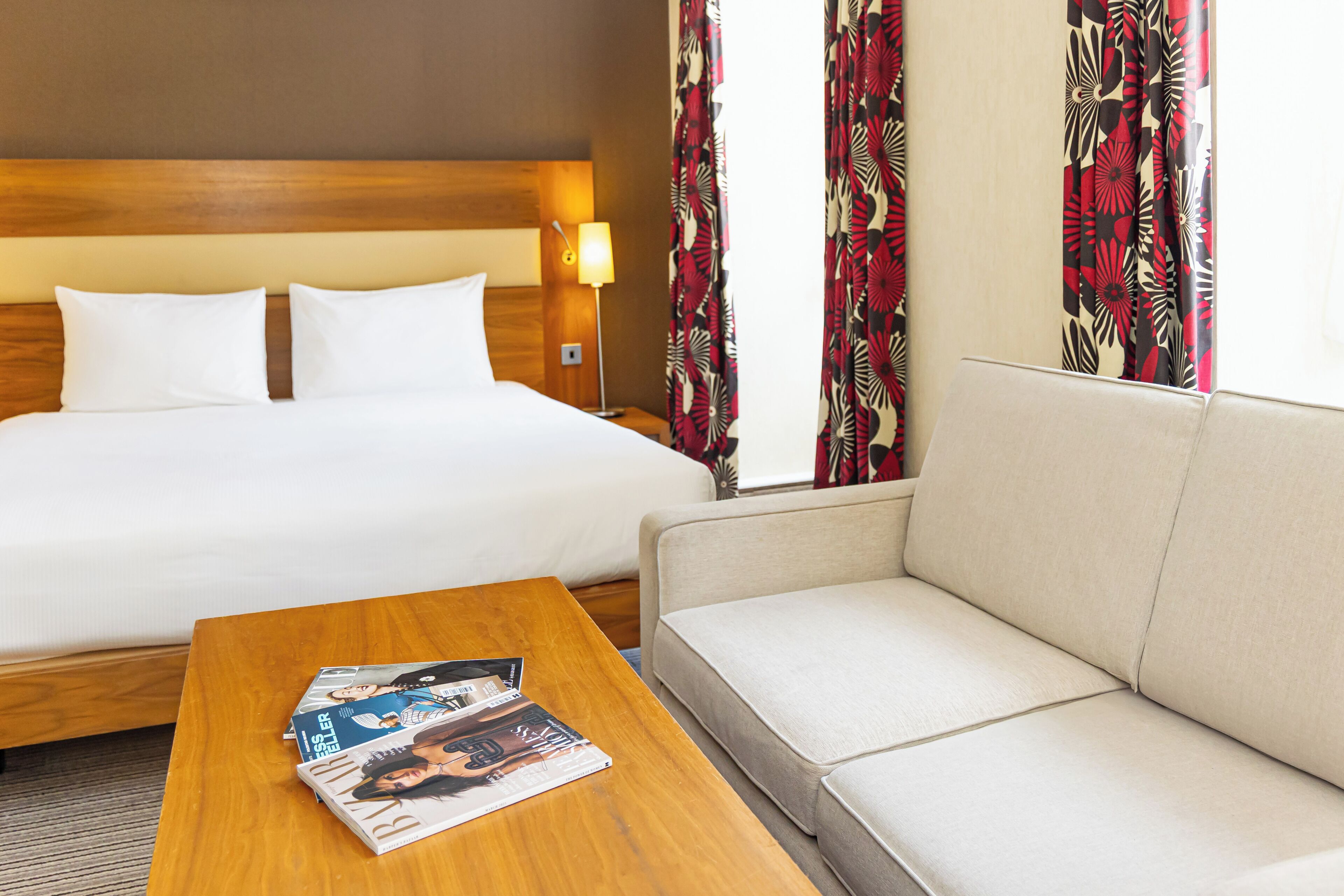 A hotel room with a bed, a sofa, a wooden coffee table with magazines, and a lamp.
