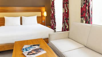A hotel room with a bed, a sofa, a wooden coffee table with magazines, and a lamp.