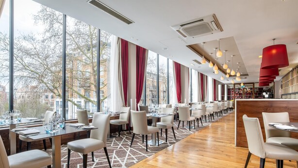 A modern restaurant with long tables, chairs, and large windows offering a view of the city. - Hilton London Olympia (London)