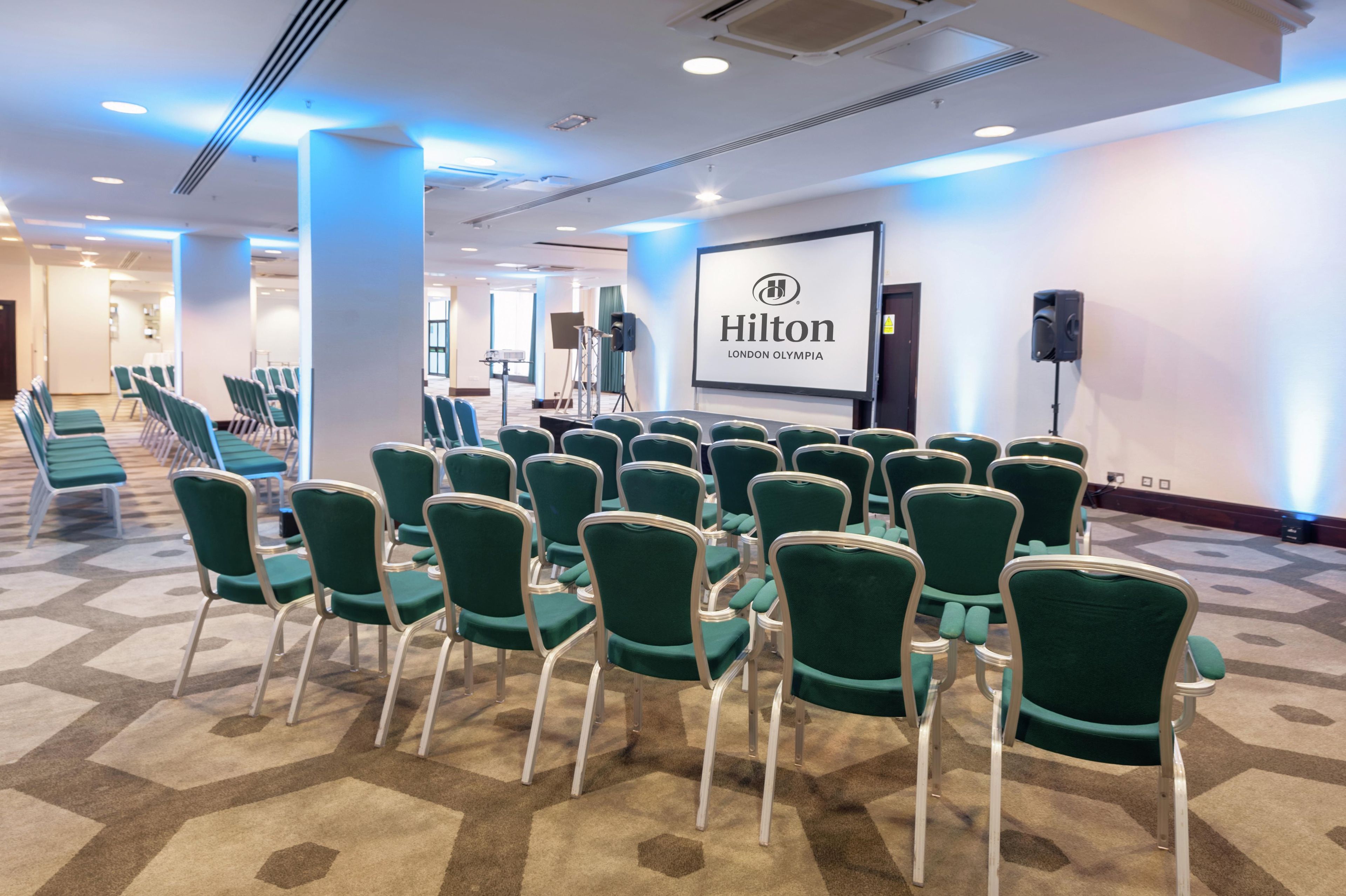 A conference room with green chairs arranged in rows, a projector screen with the Hilton logo, and a speaker on a stand.
