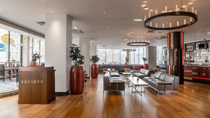 A modern lobby with a wooden reception desk labeled 'SOCIETY,' leather seating, large potted plants, and a kitchen area. - Hilton London Olympia (London)