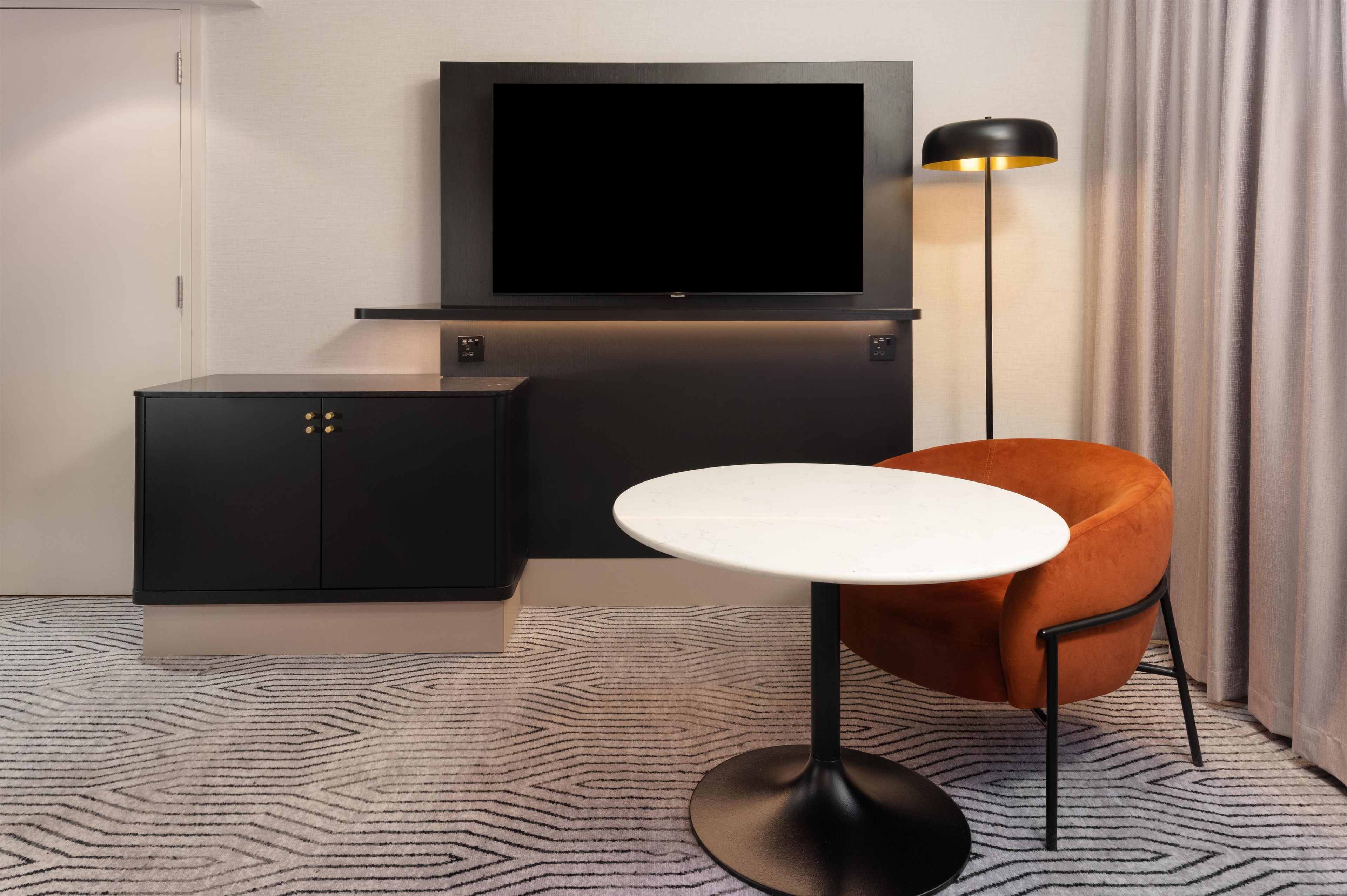 Une chambre d’hôtel moderne avec un téléviseur à écran plat fixé sur un mur sombre, une petite commode noire avec des poignées dorées, une table ronde blanche et une chaise orange avec un cadre en métal noir.