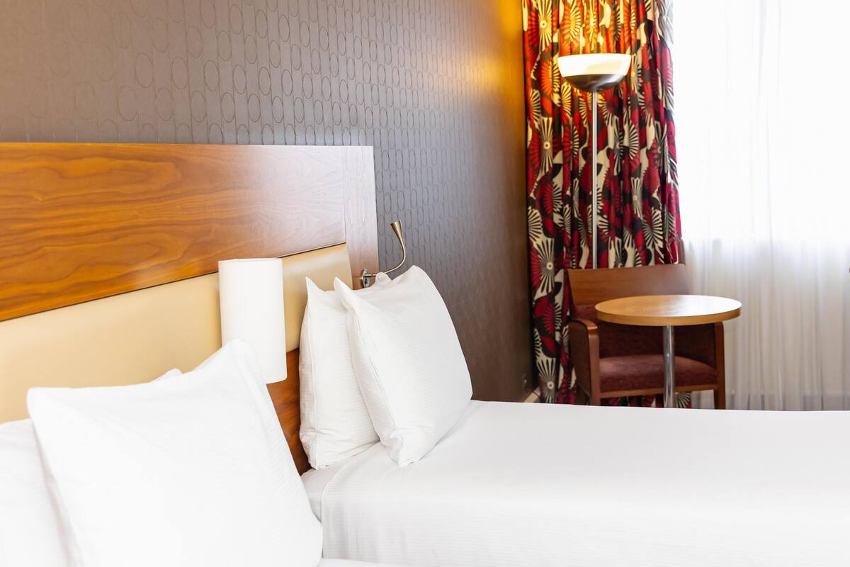 A hotel room with a wooden headboard, a bed with white linens, a round wooden table, and a patterned curtain.