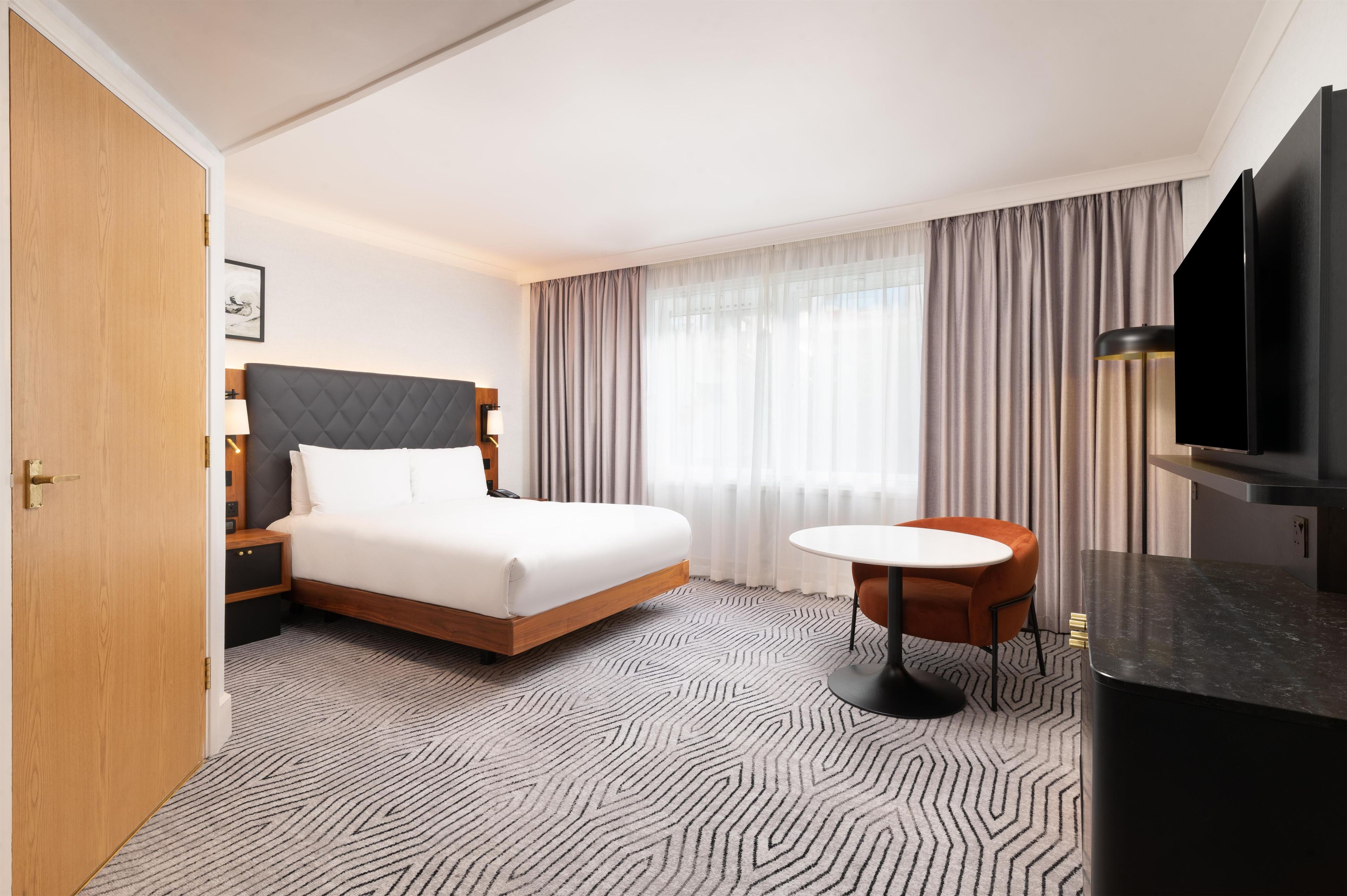 A hotel room with a bed, bedside table, lamp, TV, and a small table with a chair.