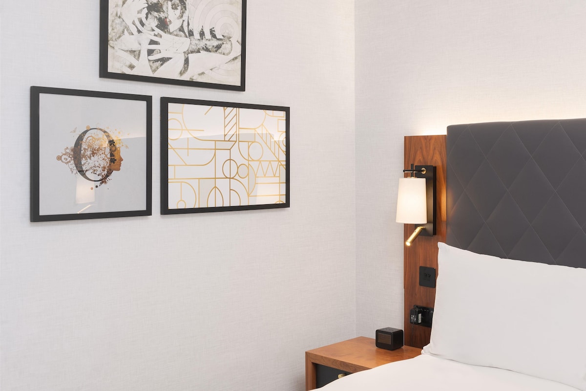 A hotel room with a bed, a nightstand with a lamp, and three framed artworks on the wall.