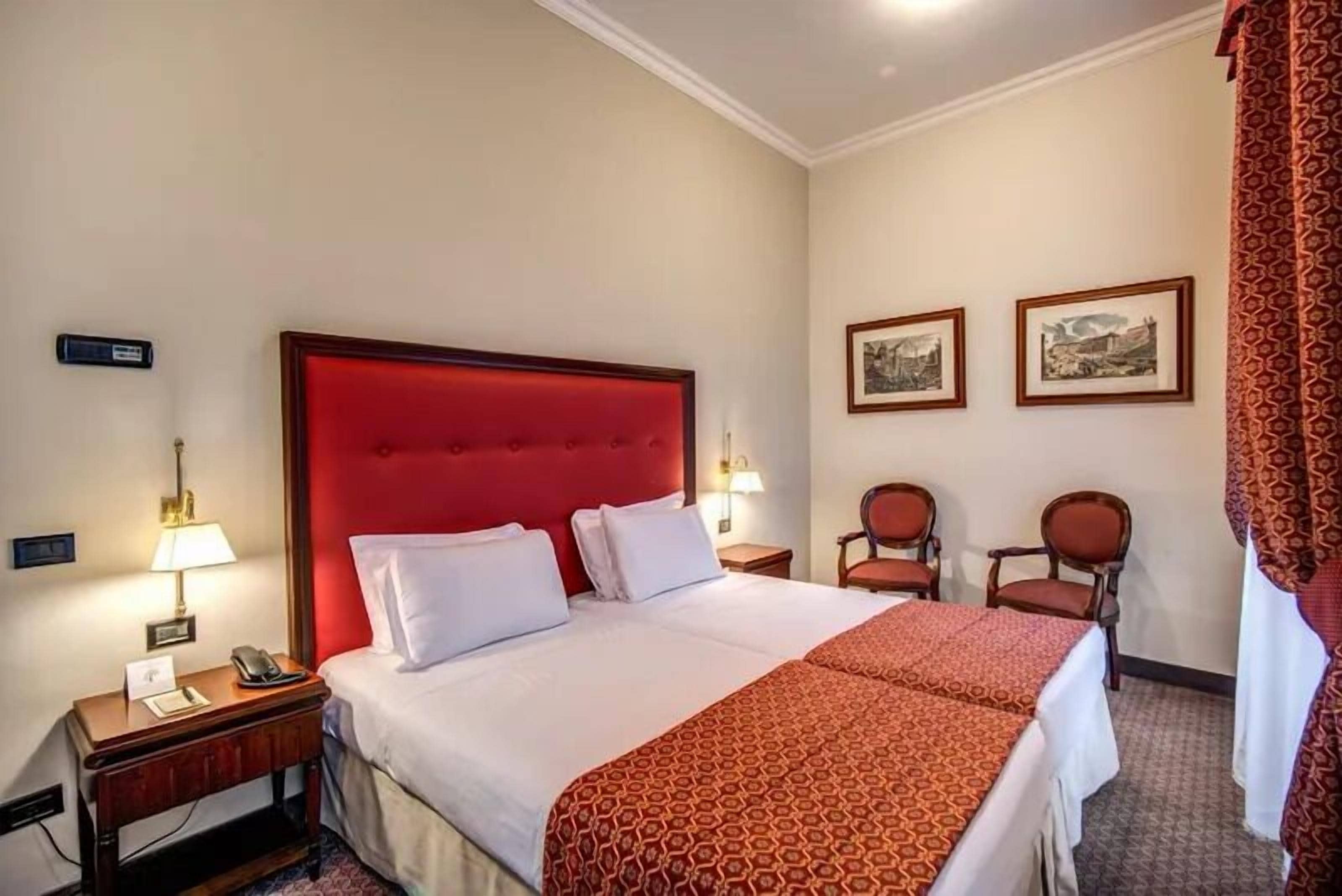 A hotel room with a large bed, a bedside table with a lamp, two chairs, and two framed pictures on the wall.