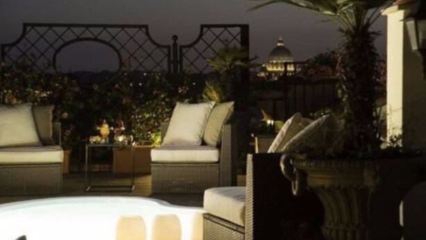 Un área de lounge en la azotea con mobiliario de mimbre, una piscina y vista a un edificio con cúpula.