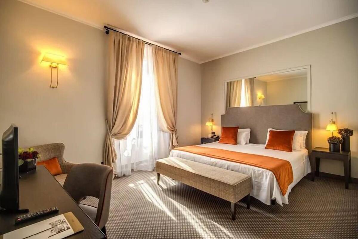 A hotel room with a bed, bedside lamps, a desk with a computer, and a large window with curtains.
