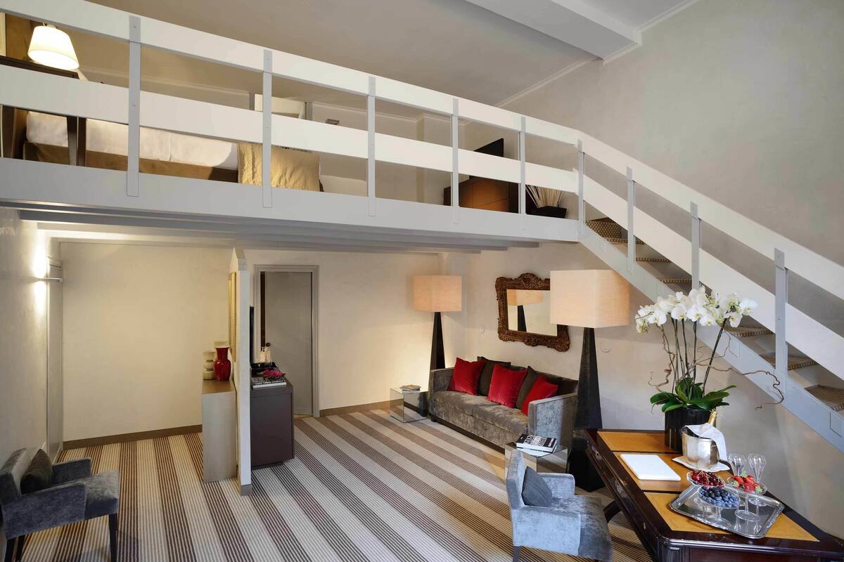 A spacious living area with a high ceiling, a staircase, a sofa set, a dining table, and a decorative plant.