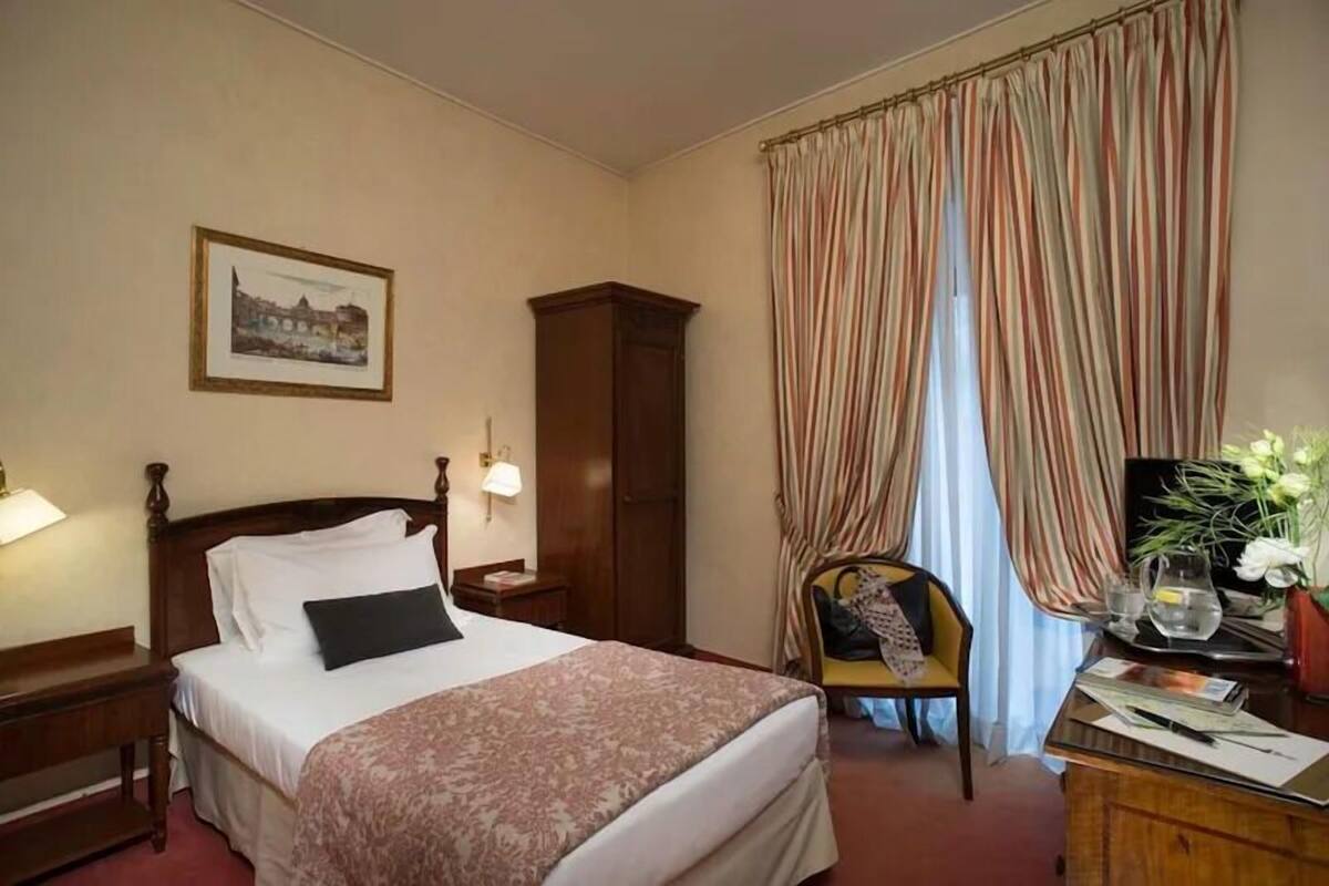 A hotel room with a bed, a wooden wardrobe, a desk with a lamp, a chair, and a window with curtains.