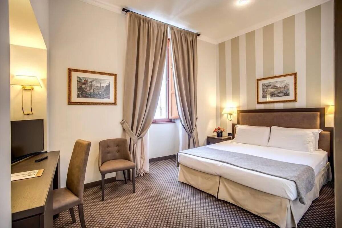 A hotel room with a bed, a desk with a chair, a TV, and two framed pictures on the walls.