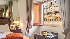 Habitación de hotel con cama, mesita de noche con lámpara, ventana con cortinas y vista a un edificio con reloj.