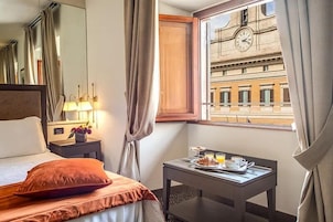 A hotel room with a bed, a bedside table with a lamp, a window with curtains, and a view of a building with a clock.