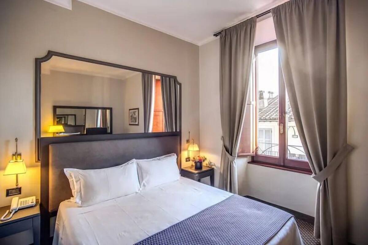 A hotel room with a large bed, a bedside table with a lamp, a telephone, and a window with curtains.
