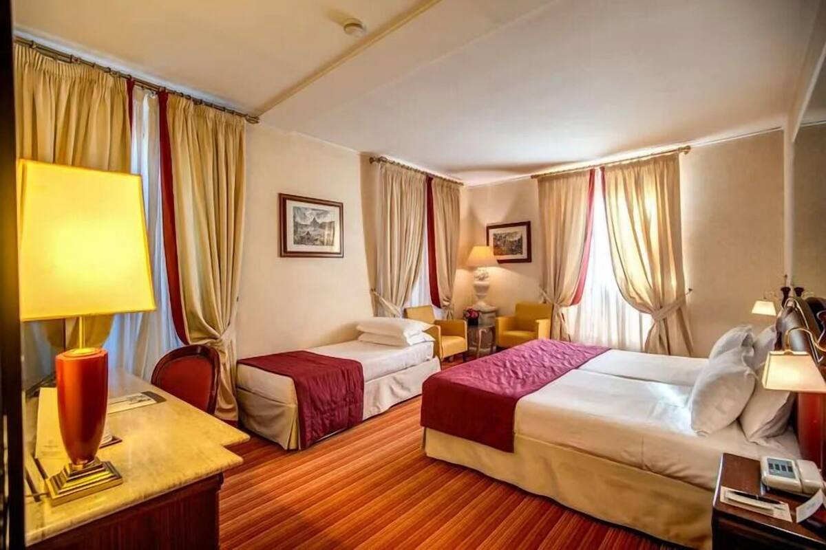 A hotel room with two beds, a desk with a lamp, a chair, and a small table with a lamp.