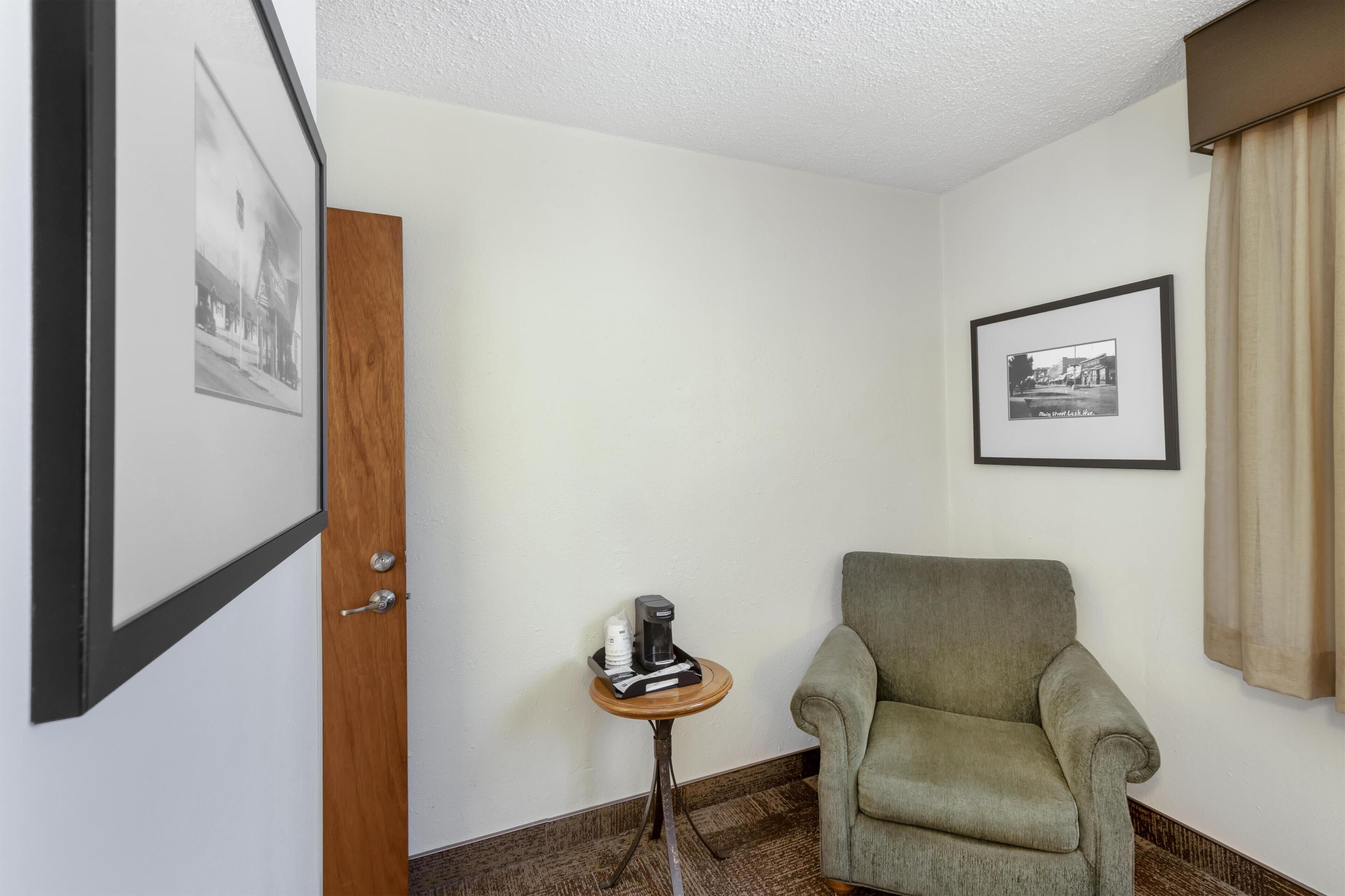 A room with a wooden door, a framed picture on the wall, a small table with a coffee maker, and a green armchair.