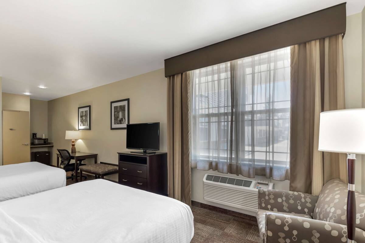 A hotel room with two beds, a desk with a chair, a television, a sofa, and a large window with curtains.