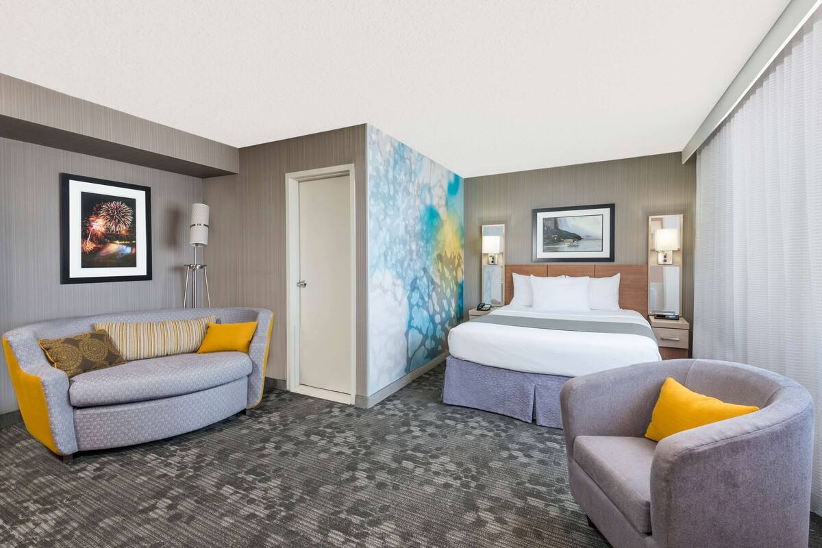 A hotel room with a bed, a sofa, a chair, a nightstand, and a wall with a framed picture.