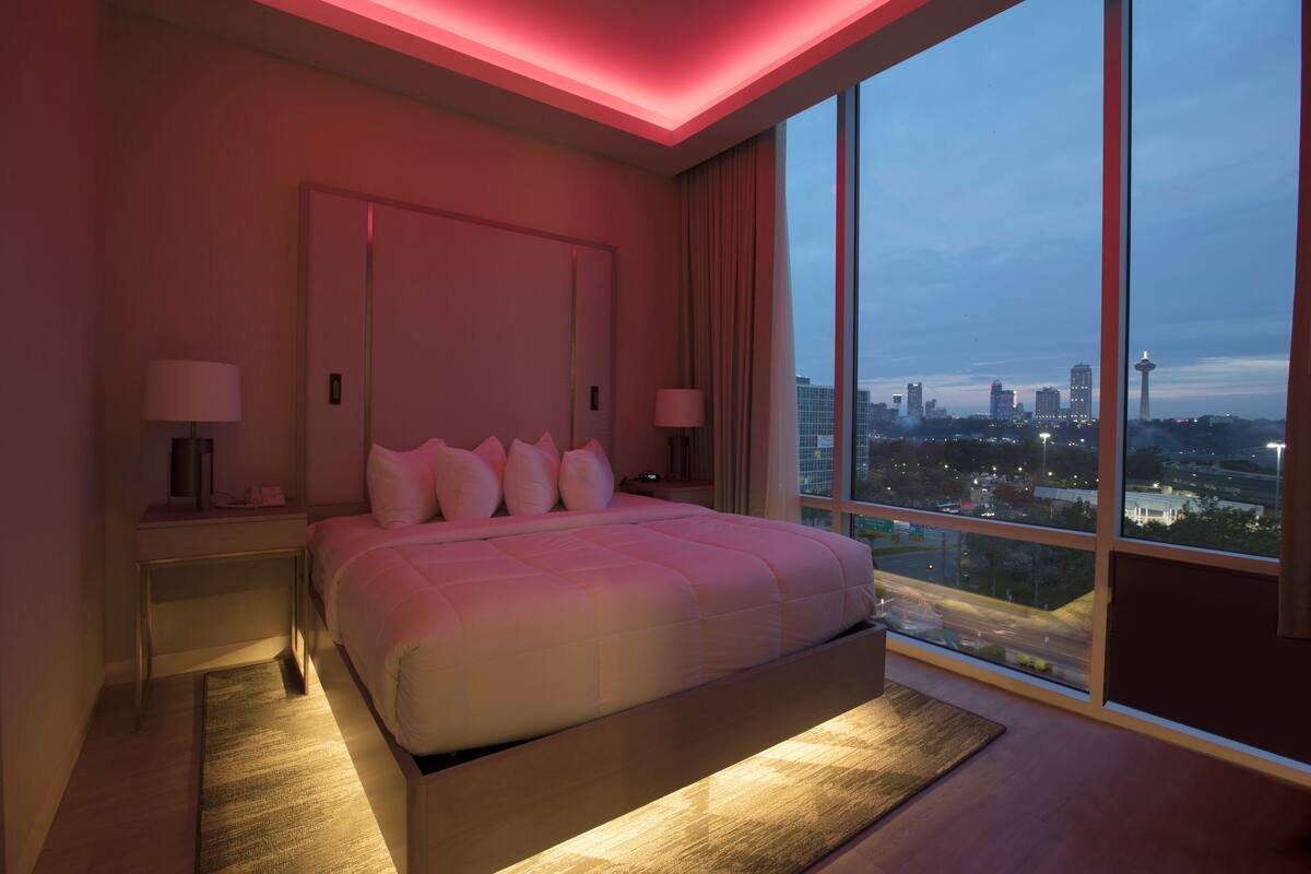 A modern hotel room with a large bed, two bedside tables with lamps, a view of the cityscape through large windows, and a city skyline visible outside.