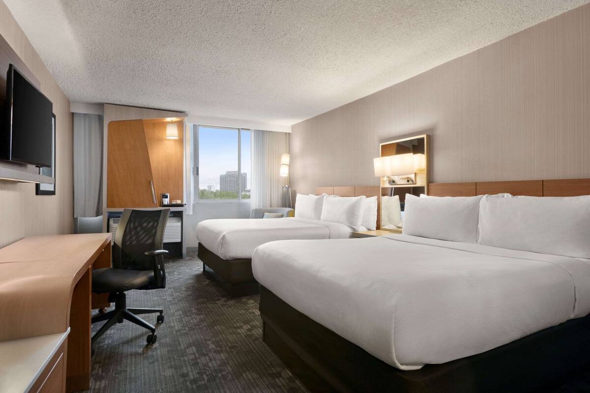 A hotel room with two beds, a desk, a chair, a TV, and a large window with a city view.